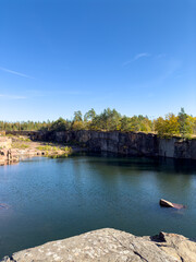 Fototapeta premium Flooded Stone Quarry Lake Surrounded by Steep Rock Cliffs. A scenic view of a deep flooded stone quarry lake enclosed by rugged rock cliffs and autumn trees under a clear blue sky.