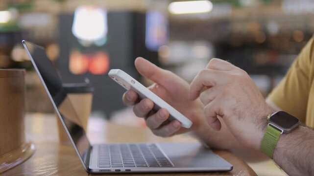 Person multitasks using phone and laptop at cafe. Hand holds phone, taps screen. Wears smartwatch, yellow shirt. Focuses on work or social media. Suitable for tech, productivity