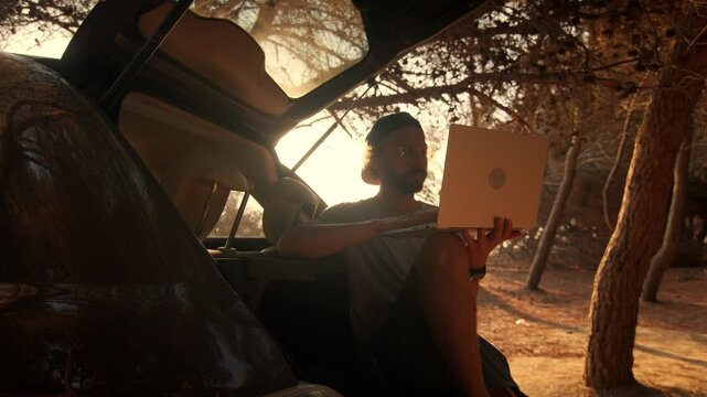 Man holding laptop inside open car trunk shot from vehicle interior, warm golden backlight and pine trees, handheld close-up.