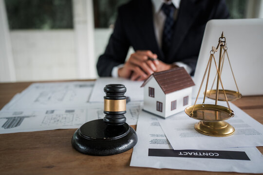 Real estate professional reviewing property documents at a desk with a house model, legal gavel, architectural plans, laptop, and scales, representing property law and transactions.