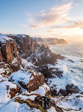 Winter sunset paints sky with golden hues over rugged, snow-dusted cliffs and a churning sea with powerful waves crashing on rocky shores.