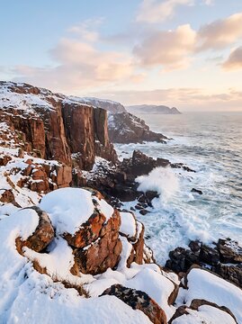 Dramatic winter sunset over a snow covered rocky coastline with powerful waves crashing against sheer cliffs under a colorful sky.