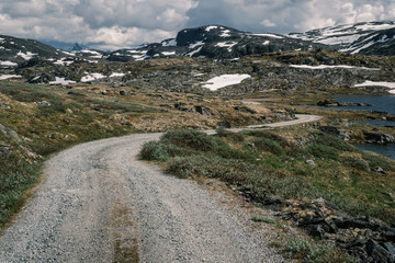 Winding gravel road carving through rocky arctic tundra slopes towards snow-covered mountains on Sognefjellet pass in Jotunheimen region, Norway © Michele