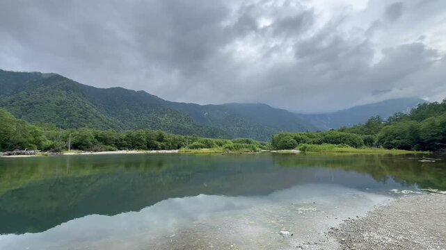 Wide static landscape shot capturing a peaceful alpine pond with lush green mountains and moody overcast sky reflecting perfectly on the calm water in Kamikochi Nagano Japan broll