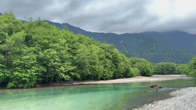 Wide static shot capturing the pristine clear water of Azusa River flowing through a lush green forest with a mountain range under a moody sky in Kamikochi Nagano Japan broll