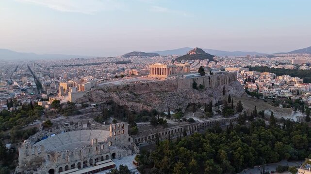 Acropolis of Athens with the Parthenon and Odeon of Herodes Atticus stands above dense city neighborhoods at twilight in a smooth drone orbit.