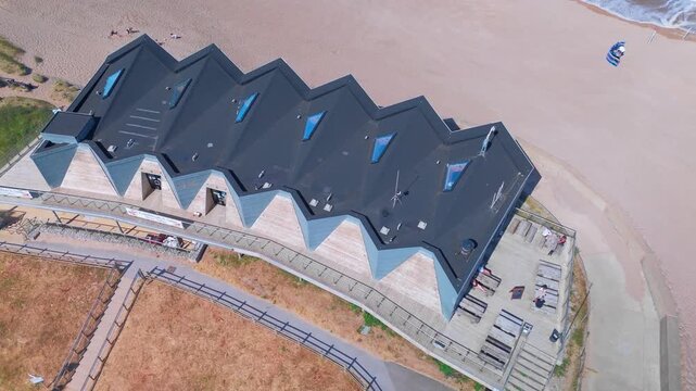 Curved roofline hugs sandy beach edge. Dark panels reflect sky above. People stroll near structure's base. Modern design blends with coastal scenery. Lincolnshire coast