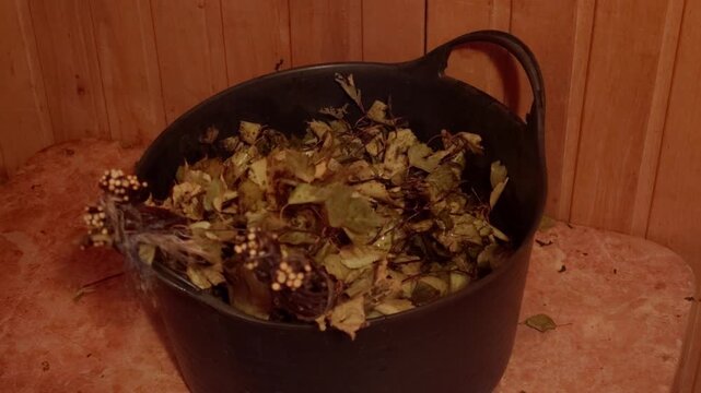 Static close up shot of black plastic container filled with soaked birch tree leaves and twigs used for traditional bathhouse whisking procedure inside timber sauna room.