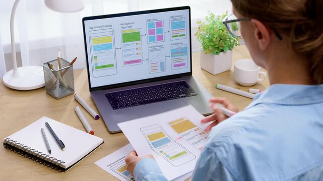 A female UX designer holds printed application wireframe sketches while comparing them with laptop screen graphics. The specialist analyzes mobile app interface layouts sitting at a work desk.