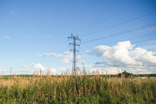 High voltage power line tower in open field under blue sky with clouds. Industrial infrastructure for electricity transmission and energy.