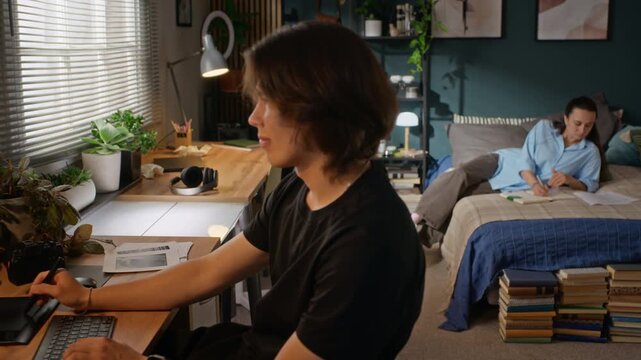Asian young man drawing on graphics tablet while Caucasian young girl studying on bed, camera panning. Ideal for remote learning or student lifestyle campaigns