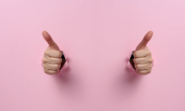 Two hands make a thumbs up gesture of approval through torn holes in pink paper.