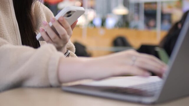 Person multitasks using phone and laptop at cafe. Hands type on keyboard and hold phone. Scene shows modern work habits in public space. Ideal for content about digital productivity or remote work
