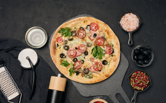 Whole pizza on a wooden board surrounded by tomatoes, olives, herbs, spices and kitchen tools
