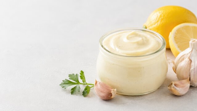 Creamy garlic aioli in a glass jar with lemons, garlic cloves and parsley on a light surface