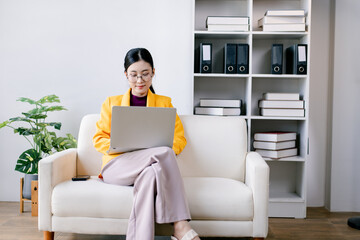 Asian businesswoman wearing glasses and a yellow blazer comfortably working on a laptop computer, managing online business from a modern home office with a bookshelf © Suphakant