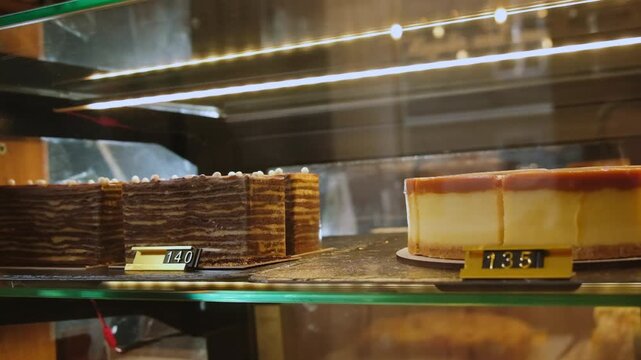 Chocolate cake and cheesecake rest behind glass display case. Dessert pieces stand beside price labels as warm cafe lights reflect across shelf