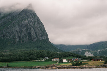 Obraz premium Coastal village with houses beneath a steep granite mountain peak shrouded in low clouds along the Helgelandskysten ferry route in Nordland, Norway.