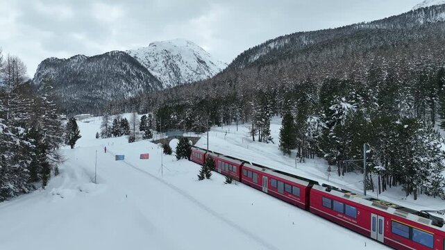 Aerial view of the Bernina Express train traveling through a snow-covered landscape with pine trees and mountains in Pontresina, Grisons, Switzerland.