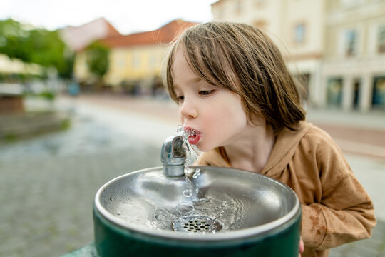 Playful young boy having fun with a public drinking fountain in the city. Laughing child splashes with fresh water outdoors during a playful urban summer moment.