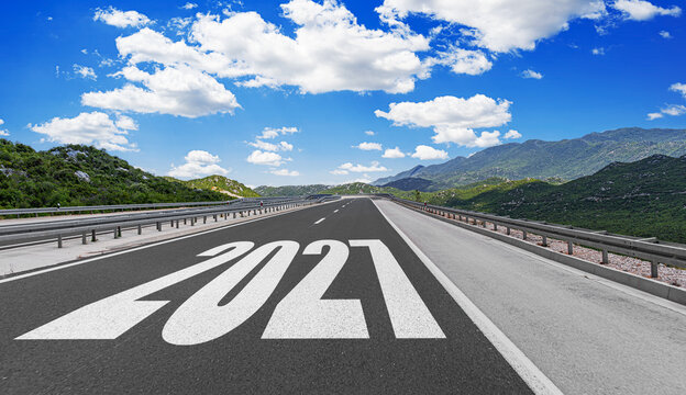 An empty asphalt highway stretches toward distant green mountains under a bright blue sky with white clouds, featuring the year 2027 painted in large white numbers on the road lane.