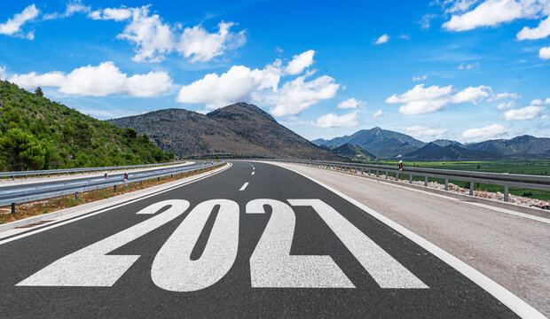 An empty open highway with the year 2027 painted on the asphalt leads toward distant mountains under a bright blue sky with white clouds.