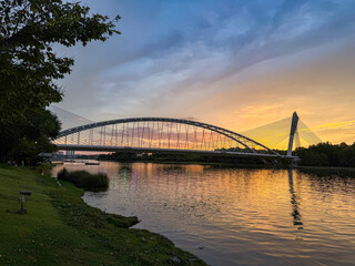 Naklejka premium Putrajaya, Malaysia. July 26, 2026. A stunning sunset view over a tranquil river, featuring a modern arched bridge illuminated by the vibrant golden and blue evening sky.