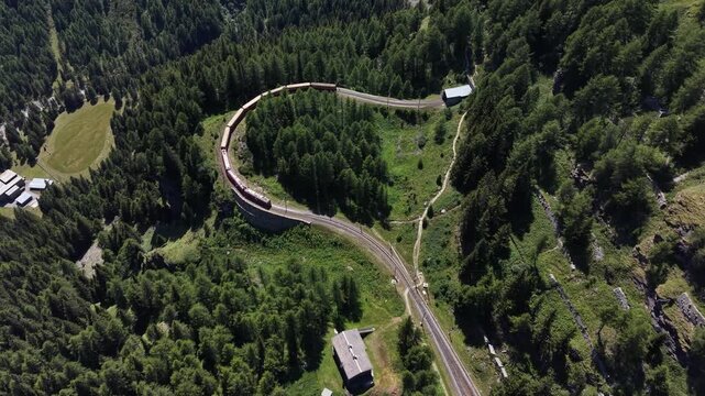 Aerial view of the Bernina Express train traveling along a curved track through a dense forest in the mountains of Poschiavo, Grisons, Switzerland.