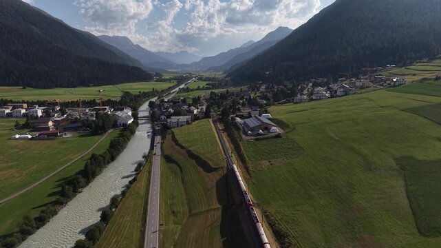 Aerial view of the Bernina Express train traveling through the lush green valley near the river in La Punt Chamues-ch, Grisons, Switzerland.