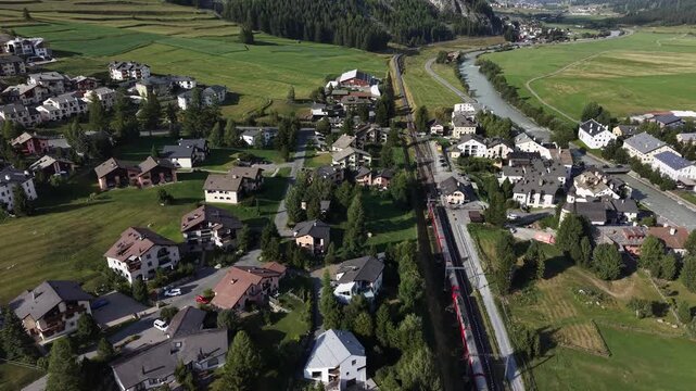 Aerial view of the Bernina Express train passing through the village of La Punt Chamues-ch surrounded by green alpine meadows and mountains in La Punt Chamues-ch, Grisons, Switzerland.