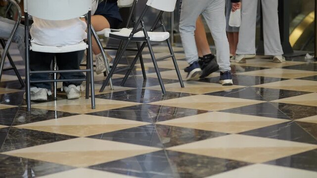 Low angle view of checkered marble floor. Black and beige tiles form a diamond pattern. People walk past folding chairs. Visible legs and feet show movement. Scene captures a busy public area