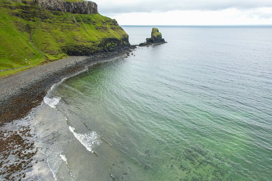 Aerial view of Talisker Bay with its rugged coastline, dark pebble beach, green grassy cliffs, and a sea stack rising from clear coastal waters Talisker, Scotland, United Kingdom.