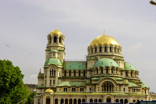 The St. Alexander Nevsky Cathedral in Sofia, Bulgaria, with its iconic golden domes and green roofs, stands majestically against a cloudy sky. 