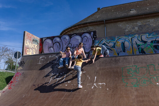 Older girls help a young girl climb a skatepark ramp outdoors