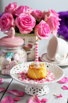 Muffin with lighted birthday candle, glass of marshmallows, cup and pink roses on table