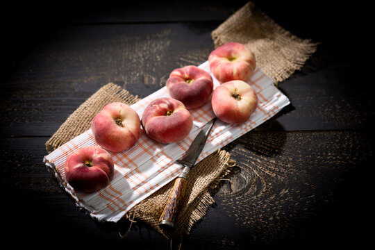 Six doughnut peaches, Prunus persica var. platycarpa, and a knife on cloth and dark wooden table