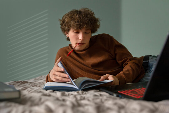 Teenage boy reads a textbook while lying on a bed at home