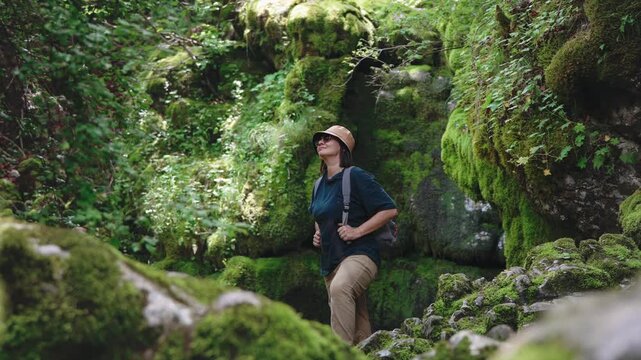 Female traveler with a backpack exploring a mysterious cave and enjoying the view while standing among large moss-covered rocks, capturing adventure, discovery and connection with wild nature.