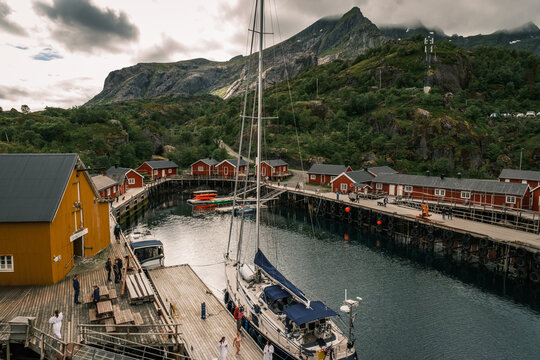 High-angle vista of Nusfjord harbour with docked sailboat, yellow cod liver oil factory, and red rorbu cabins in Lofoten, Nordland, Norway.