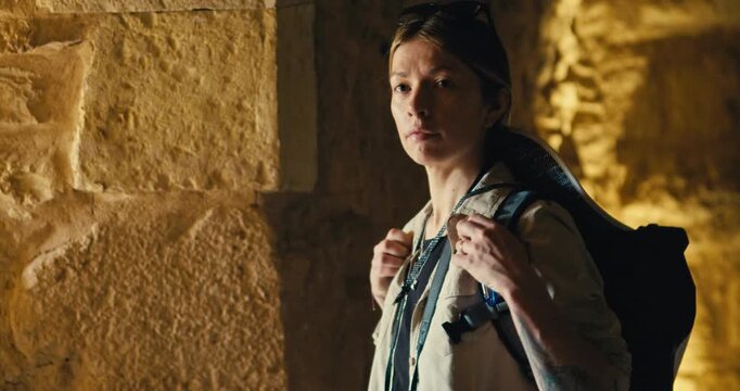 Female tourist with a backpack briefly looks toward the camera while exploring the historic stone interior of Ajloun Castle, Jordan, illuminated by warm natural light in the medieval fortress