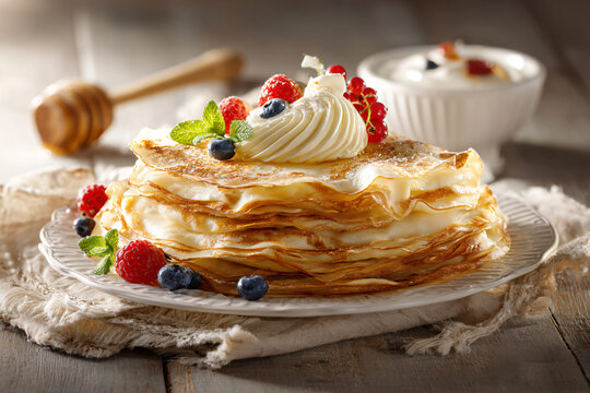 Stack of thin homemade crepes served with whipped cream, fresh raspberries, blueberries, mint and honey