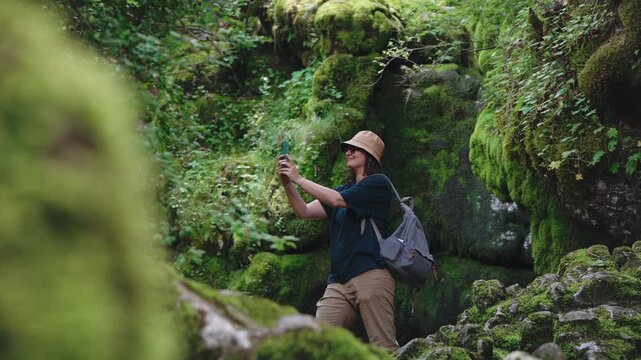 Female traveler with a backpack filming a video on her smartphone while exploring a cave among large moss-covered rocks, capturing adventure, travel content creation and discovery of wild nature.