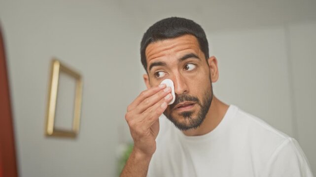 Man dabs a cotton pad to his eye and wipes cheek while inspecting skin at a vanity mirror inside a building; calm grooming.