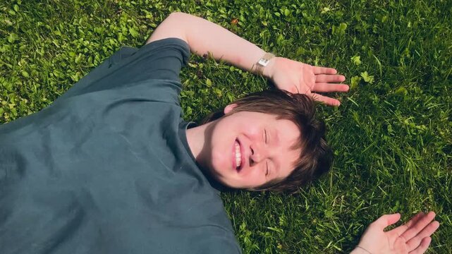 Young person lying on lush green grass in bright sunlight, eyes closed and smiling softly, viewed from overhead with one arm above head and relaxed pose in summer setting