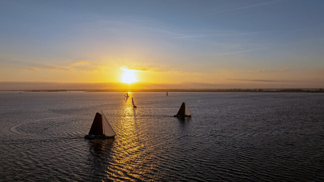 Aerial view of traditional Skutsjessilen sailing boats gliding across the water during a golden sunset in Heeg, Friesland, Netherlands.