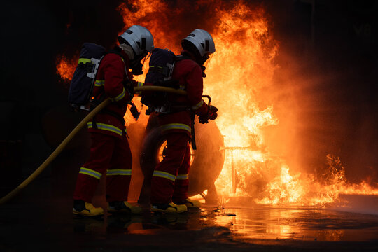 Firefighters in protective gear operate a hose near intense flames