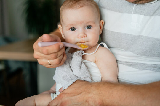 baby being fed with a plastic spoon in a calm indoor setting with white and gray tones, close-up focusing on the babys face, family life and parenting context.