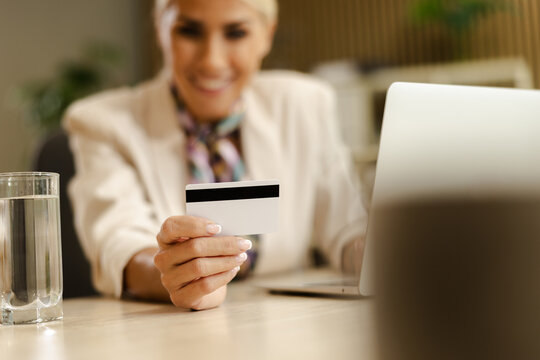 Woman at Small Office Holding a Card While Sitting at a Desk With a Laptop and a Glass of Water