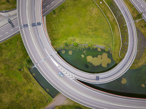 Aerial view of a highway interchange with a single car driving on the curved road surrounded by green grass and a small pond in Gothenburg, Vastra Gotaland County, Sweden.