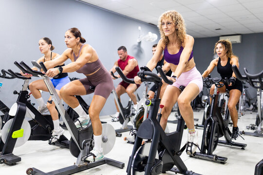 Group of people participating in an indoor cycling class at a modern fitness gym. Men and women exercising on stationary bikes during a high-intensity cardio workout.